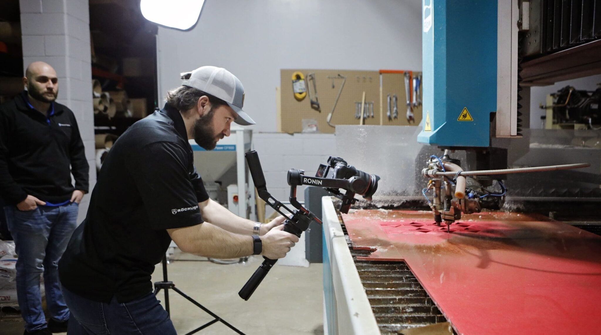 A Howell Studios camera operator films a cutting table from a gimbal while it cuts a red plastic sheet on a shop floor